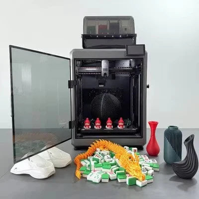 A desktop 3D printer with its door open, shown printing several objects including a white shoe, a green-and-white mahjong tile pile, a red vase, and a black bird-shaped object. The printer sits on a gray surface against a light gray background.