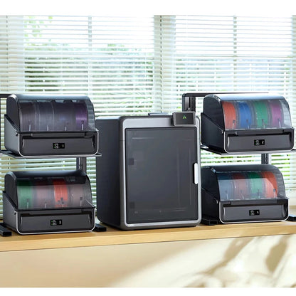 A set of three black and gray 3D printer filament storage cabinets with transparent lids showing colorful filament spools, arranged on a wooden shelf in front of window blinds.